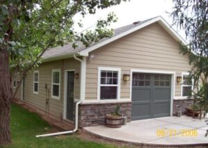 A tan two-story residential garage with a stone veneer base, featuring a grey carriage-style door and white-trimmed windows