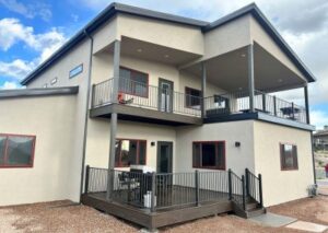 The rear view of a modern tan home featuring two levels of dark wood decks with black metal railings and red-trimmed windows